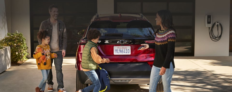 Imagem de uma família. Ao lado de um Chevrolet elétrico Bolt EUV, vermelho, pai e filha criança indo em direção ao porta malas. Atrás do carro, filho - ainda criança - mencionando abrir o porta malas do Bolt EUV. A sua frente, a mãe segurando um celular e sorrindo para o marido. Arte divulgando os seguros Chevrolet.