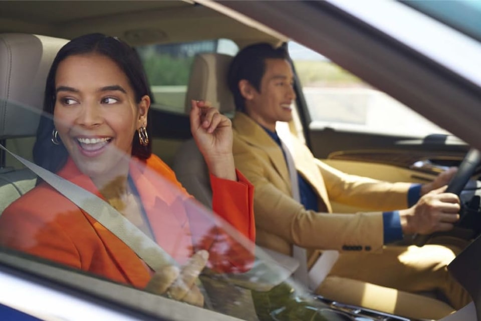 Imagem de um casal dentro de um Chevrolet. No banco do passageiro, uma mulher, preta, com cabelos lisos e longo, trajando um blazer na cor laranja, olhando para a rua e sorrindo., dirigindo e com as mãos sobre o volante. No motorista, um homem asiático, de cabelos curtos, trajando um blazer bege com as mãos no volante e sorrindo.